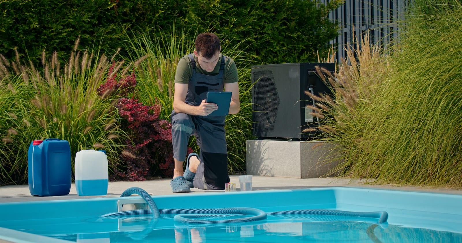 Pool service business technician reviewing route data on a tablet beside a residential pool, with chemical containers at poolside